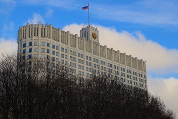 The White House of the Government of the Russian Federation with the Russian flag on the roof - against a blue sky with white clouds and tree branches in the foreground.