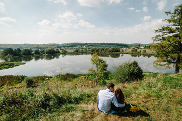 Naklejka na ściany i meble Loving couple sitting by the lake and admiring the scenery. Happy woman and man kissing on nature. Family hugging together on a sunny summer day. Back view.