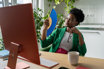 African American woman uses hand fan sits at desk with computer due to lack air conditioning system, suffers from heat. Black girl look at monitor reading industry business reports on Internet sites