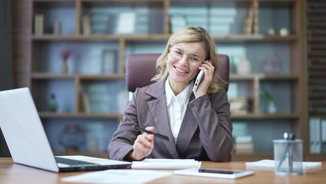 Satisfied mature businesswoman talking on the phone while sitting at the desk at the workplace in the office. Happy smiling woman in suit telling friend good news, bragging about good business results