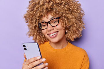 Happy beautiful European woman with curly hair holds mobile phone reads funny message enjoys making shopping online wears casual orange jumper and spectacles isolated over purple background.