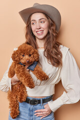 Vertical shot of cheerful woman wears cowboy hat blouse and jeans poses with small poodle puppy plays with her favorite pet looks gladfully aside isolated over brown background. Company of best friend