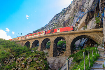 Red train passing by Devil's bridge at St. Gotthard pass on the Swiss Alps in Andermatt, Switzerland