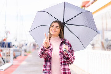 Young pretty Romanian woman holding an umbrella at outdoors smiling and showing victory sign