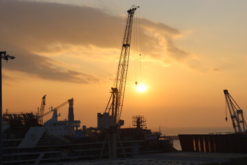 Fototapeta premium Genova, Italy - February 5, 2023: Beautiful view to the port of Genova in winter with sunset colours reflected on the water. Golden hour hidden behind the clouds and the industrial zone of the port.