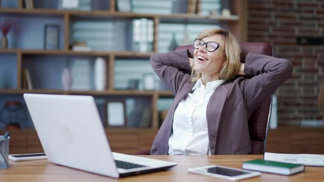 Happy mature businesswoman finished work on laptop while sitting at desk at workplace in office. A beautiful female smiles, throws her hands behind her head and stretches on a chair. Work done
