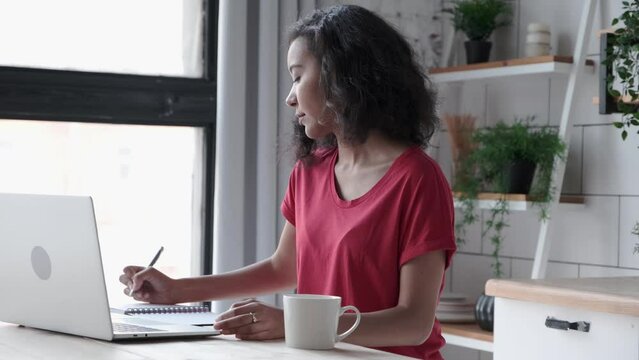 Young African American woman sits at a table in front of a laptop at home. Attractive curly woman in a red t-shirt makes notes and sketches, draws up a plan in a paper notebook, freelance work