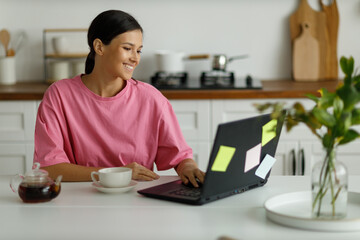Happy girl with laptop checks her mail, looks through messages or funny videos. Video communication online. Beautiful young woman works remotely from home, sitting at the dining table in the kitchen.