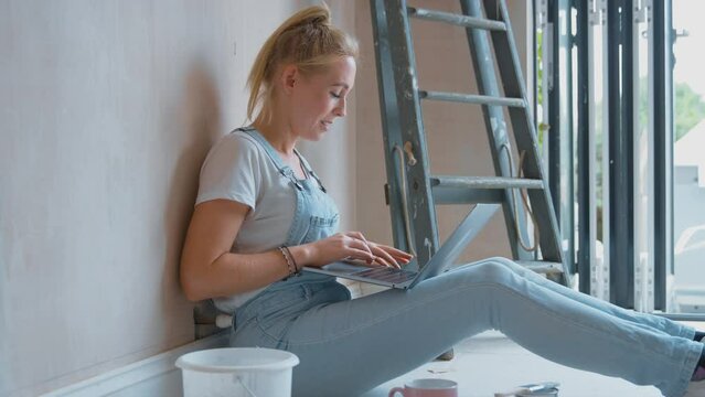 Woman renovating room at home sits on floor with laptop taking a break from decorating room - shot in slow motion