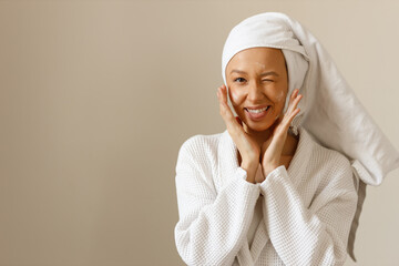 Portrait of attractive playful brunette woman in white robe and towel on her head, applying lotion...