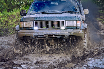Blue SUV participating in an off road race is driving through water mud, and splashes are flying in all directions. The four wheel drive car powerfully climbs up slope, smoke billows from exhaust pipe