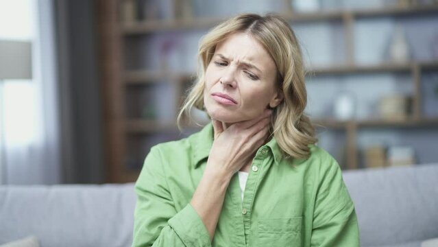 Close up of sick mature blonde woman having sore throat sitting in living room on sofa at home. Female has first symptoms of cold and flu virus, pneumonia, bronchitis and respiratory tract infection