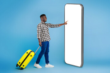 Handsome millennial black guy with bright suitcase using huge phone