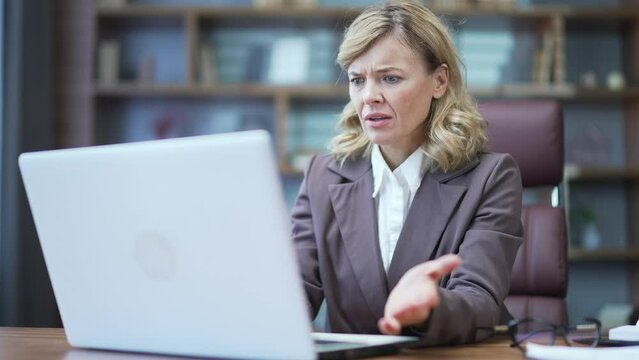 Disoriented mature businesswoman looking confusedly at laptop screen at desk at workplace in office. Sad female spreads her hands and cannot understand the mistakes in the work of the computer program
