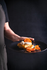 The Chef in  black  holds in his hands plate with dessert:  Belgian waffles with whipped cream and orange. Selective focus.