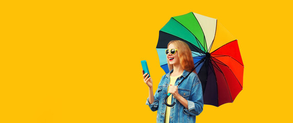 Portrait of happy smiling young woman looking at smartphone with colorful umbrella isolated on yellow background, blank copy space for advertising text