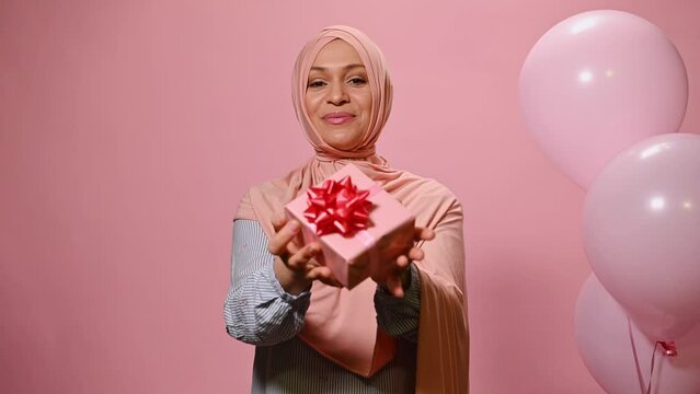 Beautiful middle-aged Arab Muslim woman with head covered in hijab, showing a cute present at camera, smiling a toothy smile at camera, posing with helium balloons, isolated on pik color background