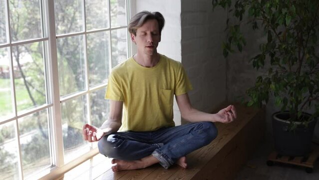 Serious young man meditating at home sitting on windowsill holding hands in mudra with closed eyes in cozy apartment. People mental healthcare and relaxation concept.