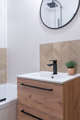 Ceramic sink with tap and wooden cabinet, round mirror and herringbone pattern on the wall in modern bathroom. Stylish interior with minimalism. Vertical.