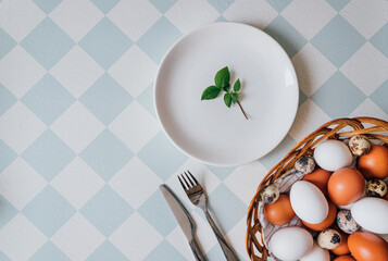top view, close-up of a spring still life for Easter, vertical frame, background of blue and white rhombuses, an empty white plate with one green leaf of a tree and a wicker basket with colorful eggs