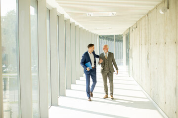 Young and a senior businessman walk down an office hallway, deep in conversation