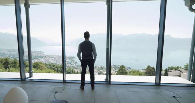 One Young Businessman Inside Modern Building Looking Through Window Overlooking Lake City Landscape View