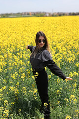 Fototapeta premium Cute young woman in yellow flower field. Her hair is blowing with wind. She has a black belt around her waist.