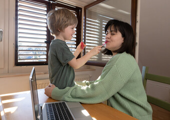 The child sits on the table next to the mother and paints her lips, work from home