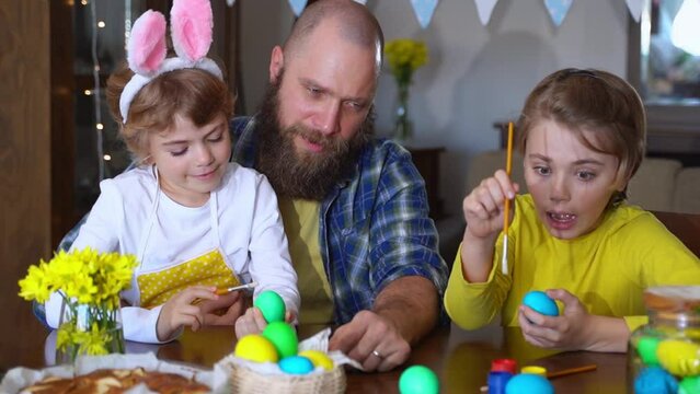 Easter Family traditions. Father and two caucasian happy children with bunny ears dye and decorate eggs with paints for holidays while sitting together at home table. Kids embrace and smile in cozy.
