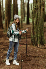 Glad millennial caucasian lady in hat with backpack, trekking sticks, resting in cold forest, enjoy adventure