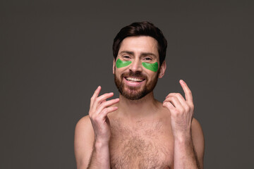 Shirtless man with hydrogel eye patches smiling at camera isolated on grey background 