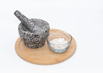 Mortar and pestle made from black and gray granite and a clear glass bowl with white pills waiting to be pulverized are on a round wooden board against a white background.
