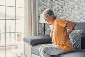 Unhappy mature woman touching back, feeling pain, sitting on couch in living room, unhealthy middle...