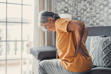 Unhappy mature woman touching back, feeling pain, sitting on couch in living room, unhealthy middle...