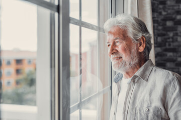 Happy thoughtful older 70s man looking out of window away with hope, thinking of good health, retirement, insurance benefits, dreaming of future. Elderly pensioner waiting meeting with family.