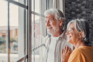 Happy bonding loving middle aged senior retired couple standing near window, looking in distance, recollecting good memories or planning common future, enjoying peaceful moment together at home.. - obrazy, fototapety, plakaty