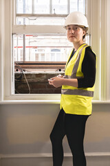 Vertical photo of a chartered civil engineer lady using a calculator in front of a window in an indoor construction site, wears hard hat, yellow personal protective equipment, looking at camera