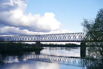 Silhouette of the railway bridge across the river, the reflection of the bridge in the river against the background of the blue sky and clouds