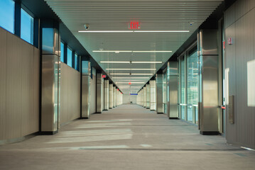 corridor of empty building with exit signs lit in red and windows on one side, during the day