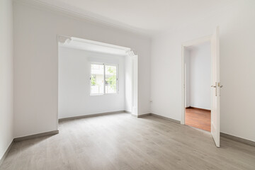 An empty spacious room, sunlight penetrates through the window during daylight hours. On floor there is wooden parquet in gray color. In open doorway is corridor to the rest of the apartment.