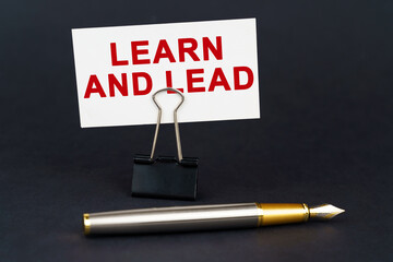 On the black surface is a pen, an office paper clip with a business card on which is written - LEARN AND LEAD