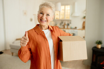 Stylish elderly female with short haircut posing with carton box showing thumb up with smiling...