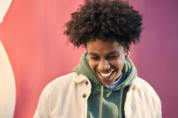 Authentic candid shot of young happy African American cool teen guy laughing on red wall lit with sunlight. Smiling cool rebel gen z teenager model standing outdoors having fun, portrait outdoors. - obrazy, fototapety, plakaty