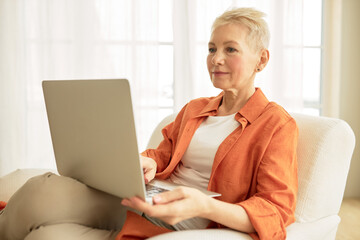Indoor portrait of cool mature woman of 60s with stylish haircut watching movie or video,playing online games, reading book or blogging sitting in armchair with laptop against big windows