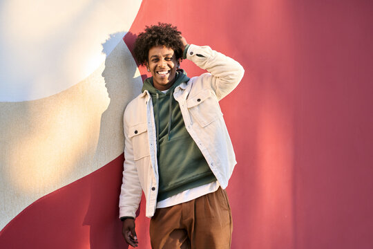 Young happy African American teen guy laughing on red wall lit with sunlight. Smiling cool ethnic generation z teenager student model standing looking at camera posing for portrait outdoors.