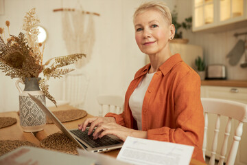 Indoor portrait of senior positive woman with short haircut using laptop to make payments online on...