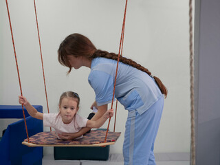 Child with physiotherapist on swing during sensory integration session. Little girl in rehabilitation centre doing exercises for development functions of the vestibular apparatus