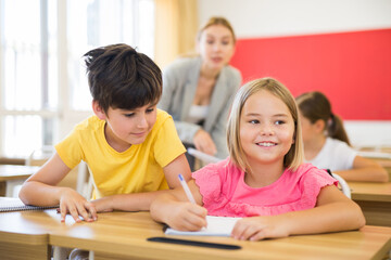 Cheerful preteen schoolchildren sitting at lesson in classroom, inquisitive boy peeking at workbook of cute smiling girl