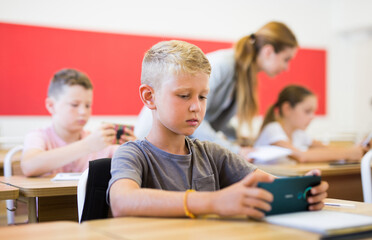 Interested absorbed preteen boy using smartphone while studying in schoolroom with classmates and...