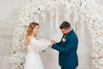 happy bride and groom exchange rings in an arch of artificial flowers.